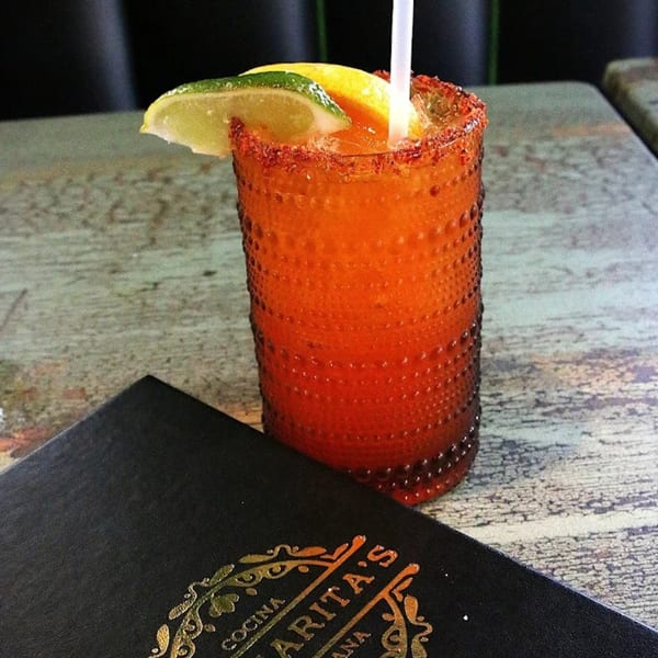 A vibrant orange drink with a chili-lime rim, garnished with lime and orange slices, next to a Margarita's Cocina Mexicana menu.