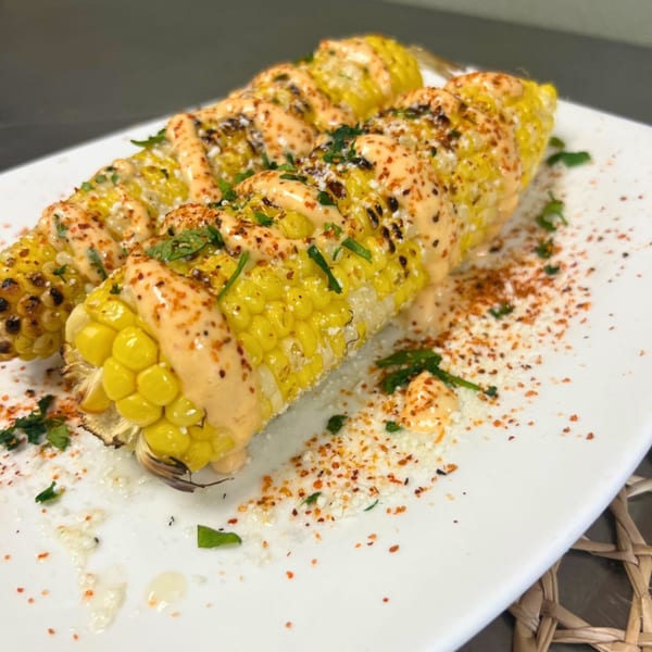 Two elotes, Mexican street corn, on a white plate, topped with creamy sauce, cheese, and chili powder.
