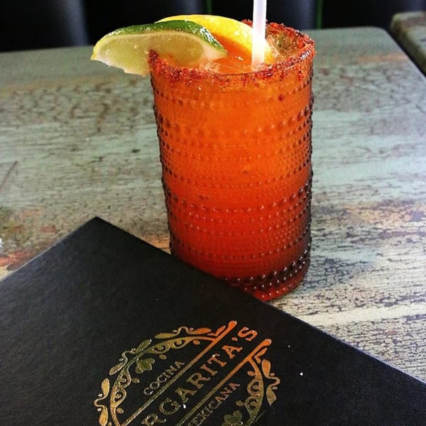 A vibrant orange drink with a chili-lime rim, garnished with lime and lemon, next to a Margarita's Cocina Mexicana menu.