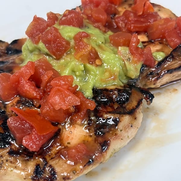 Grilled chicken breast topped with guacamole and diced tomatoes on a white plate.