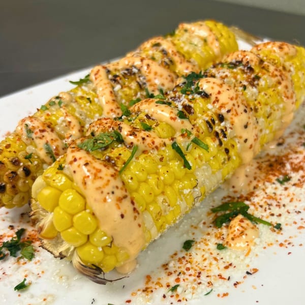 Two elotes, Mexican street corn, grilled and topped with creamy sauce, cheese, and chili powder.