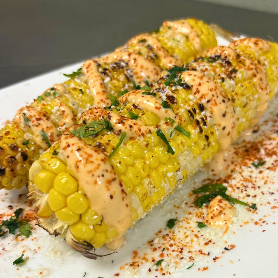 Two elotes, Mexican street corn, grilled and topped with creamy sauce, cheese, and chili powder.